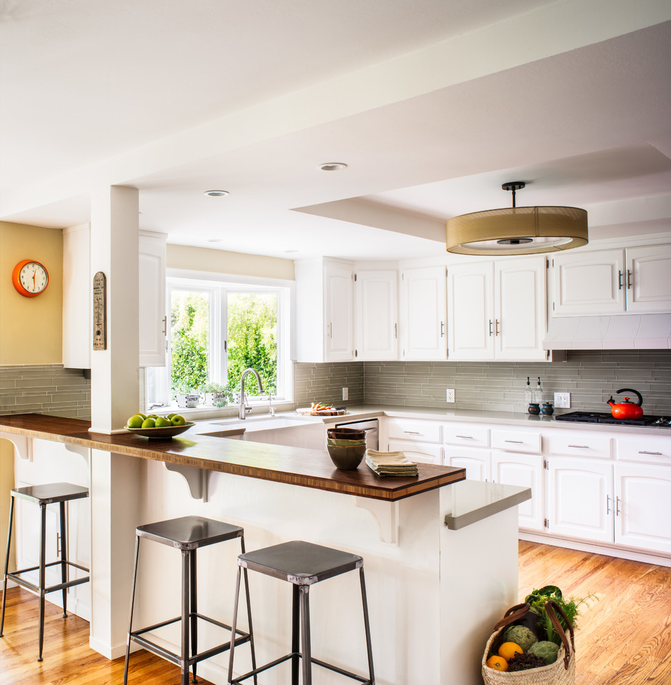 Photo of a large transitional u-shaped eat-in kitchen in San Francisco with a farmhouse sink, raised-panel cabinets, white cabinets, quartz benchtops, glass tile splashback, stainless steel appliances, light hardwood floors and grey splashback.