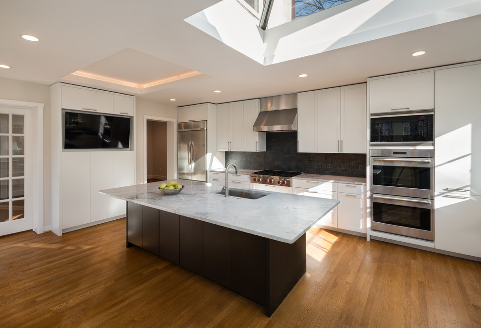 Photo of a large contemporary u-shaped separate kitchen in DC Metro with a drop-in sink, recessed-panel cabinets, white cabinets, black splashback, slate splashback, stainless steel appliances, light hardwood floors, with island, brown floor and quartzite benchtops.