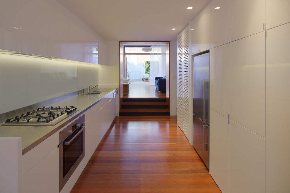 Contemporary galley eat-in kitchen in Other with an integrated sink, flat-panel cabinets, white cabinets, stainless steel benchtops, white splashback, glass sheet splashback and stainless steel appliances.