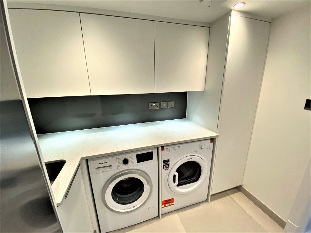 This is an example of a small l-shaped utility room in London with a drop-in sink, flat-panel cabinets, white cabinets, quartzite benchtops, brown splashback, glass sheet splashback, white walls, a side-by-side washer and dryer and white benchtop.