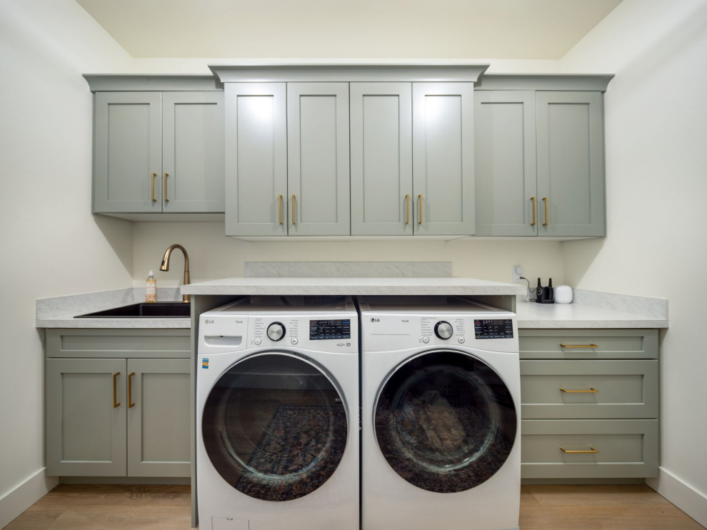 Design ideas for a large country single-wall dedicated laundry room in Other with a drop-in sink, shaker cabinets, green cabinets, laminate benchtops, white walls, light hardwood floors, a side-by-side washer and dryer, beige floor and white benchtop.