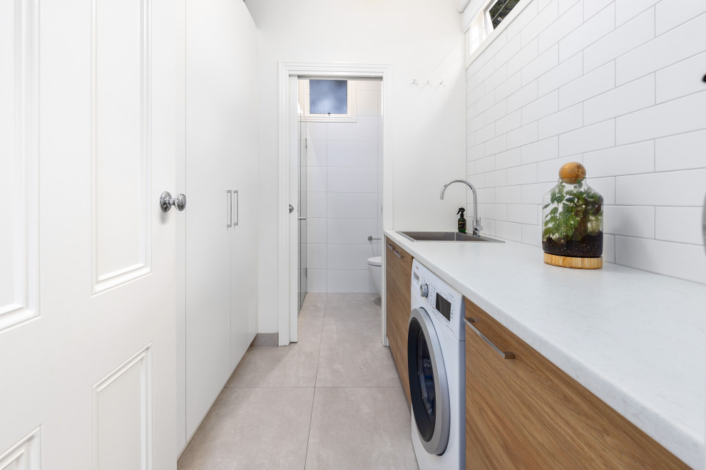 Photo of a medium sized scandinavian galley separated utility room in Melbourne with an utility sink, flat-panel cabinets, light wood cabinets, engineered stone countertops, white splashback, metro tiled splashback, white walls, porcelain flooring, a concealed washer and dryer, beige floors and white worktops.