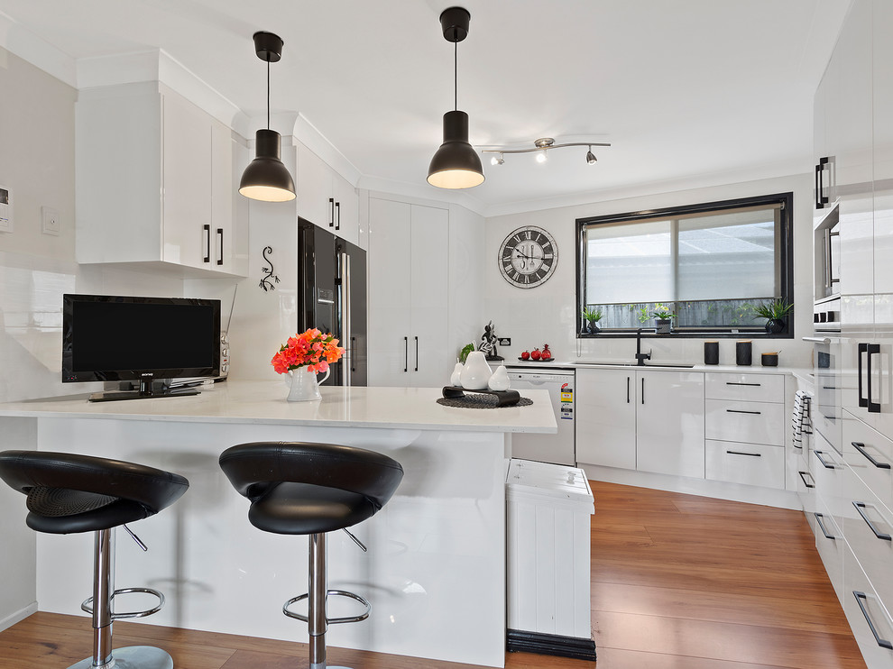 Inspiration for a mid-sized contemporary u-shaped eat-in kitchen in Gold Coast - Tweed with flat-panel cabinets, solid surface benchtops, white splashback, ceramic splashback, an undermount sink, black appliances, medium hardwood floors and a peninsula.