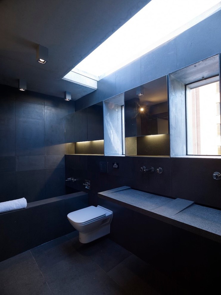 Photo of a mid-sized modern master bathroom in Sydney with flat-panel cabinets, an alcove tub, a wall-mount toilet, black tile, stone tile, black walls, porcelain floors, an integrated sink, tile benchtops, black floor and black benchtops.