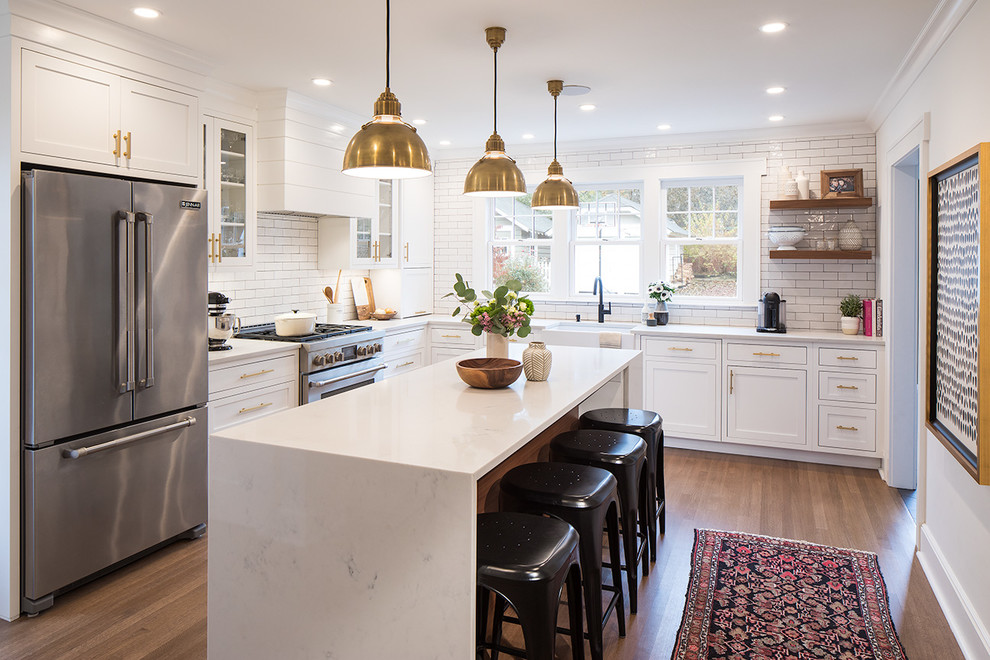 Photo of a large contemporary l-shaped eat-in kitchen in New York with a farmhouse sink, shaker cabinets, white cabinets, quartz benchtops, white splashback, ceramic splashback, stainless steel appliances, medium hardwood floors, with island and brown floor.