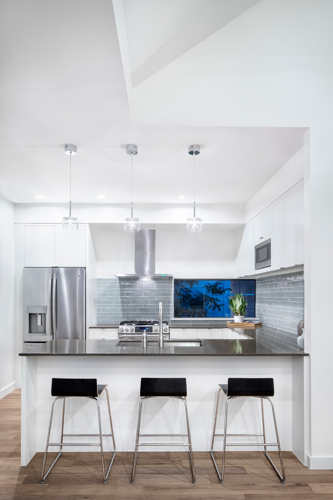 Design ideas for a mid-sized contemporary u-shaped separate kitchen in Austin with an undermount sink, flat-panel cabinets, white cabinets, grey splashback, stainless steel appliances, light hardwood floors, a peninsula, solid surface benchtops, glass tile splashback and brown floor.