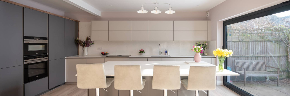 Inspiration for a large contemporary l-shaped open plan kitchen in Hertfordshire with a double-bowl sink, flat-panel cabinets, white cabinets, quartz worktops, white splashback, black appliances, an island, white worktops, exposed beams and feature lighting.