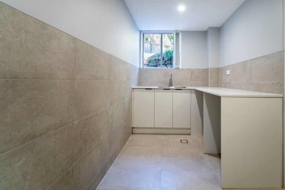 Example of a large trendy l-shaped ceramic tile and beige floor dedicated laundry room design in Sydney with a drop-in sink, flat-panel cabinets, white cabinets, quartz countertops, beige backsplash, ceramic backsplash, beige walls, a side-by-side washer/dryer and white countertops