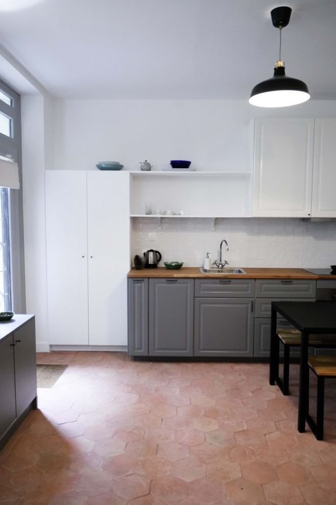 This is an example of a mid-sized transitional single-wall open plan kitchen in Paris with an undermount sink, beaded inset cabinets, grey cabinets, wood benchtops, white splashback, panelled appliances, terra-cotta floors, red floor, beige benchtop and ceramic splashback.