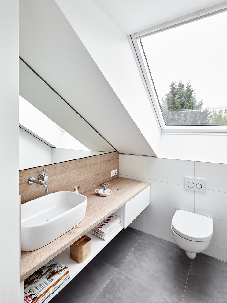 This is an example of a contemporary powder room in Dusseldorf with open cabinets, white cabinets, a wall-mount toilet, white tile, ceramic tile, white walls, a vessel sink, wood benchtops, grey floor and brown benchtops.