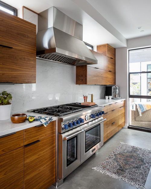 Warm minimalist kitchen cabinet design featuring walnut slab cabinets with horizontal grain, white quartz backsplash, stainless pro range and hood, black pulls, and a concrete floor