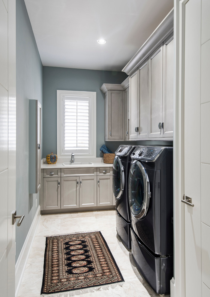 Design ideas for a mid-sized transitional l-shaped laundry room in Miami with a drop-in sink, recessed-panel cabinets, white cabinets, solid surface benchtops, grey walls, travertine floors and a side-by-side washer and dryer.