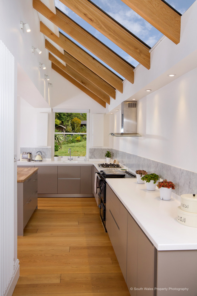 Mid-sized contemporary l-shaped open plan kitchen in Other with with island, an integrated sink, flat-panel cabinets, beige cabinets, solid surface benchtops, grey splashback, marble splashback, black appliances, medium hardwood floors, brown floor, white benchtop and exposed beam.