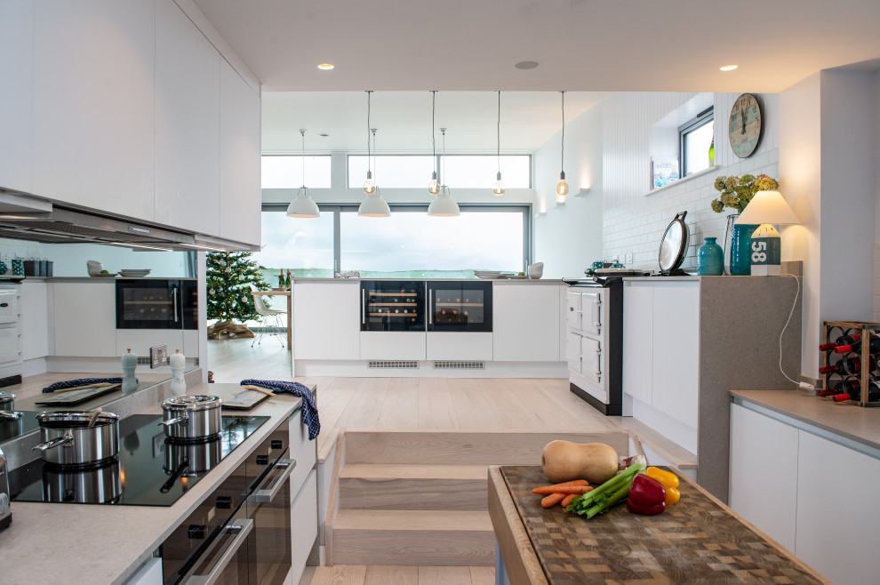 Photo of a medium sized nautical galley open plan kitchen in Cornwall with flat-panel cabinets, white cabinets, composite countertops, white splashback, cement tile splashback, black appliances and beige worktops.