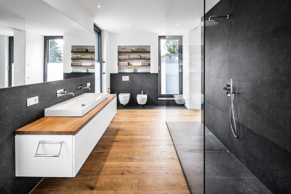 Large contemporary bathroom in Cologne with flat-panel cabinets, white cabinets, an open shower, a bidet, black tile, white walls, medium hardwood floors, a trough sink, wood benchtops, an open shower and brown benchtops.