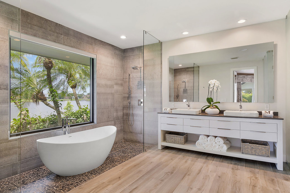 Design ideas for a beach style master bathroom in Miami with flat-panel cabinets, white cabinets, a freestanding tub, a curbless shower, gray tile, grey walls, medium hardwood floors, a drop-in sink, brown floor, an open shower and grey benchtops.