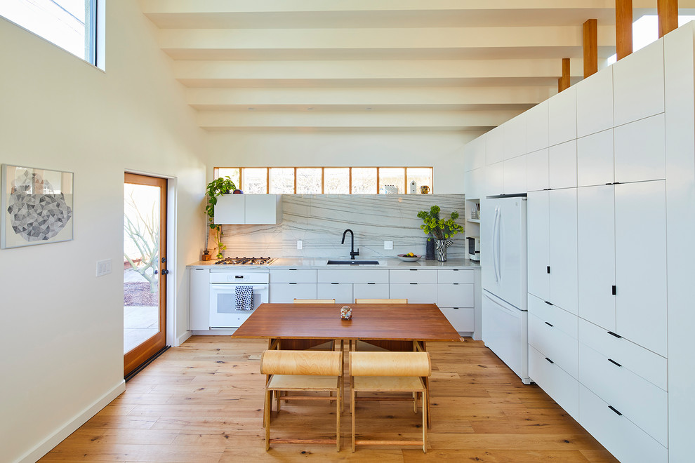 This is an example of a midcentury eat-in kitchen in Los Angeles with an undermount sink, flat-panel cabinets, white cabinets, white splashback, stone slab splashback, white appliances, medium hardwood floors and white benchtop.
