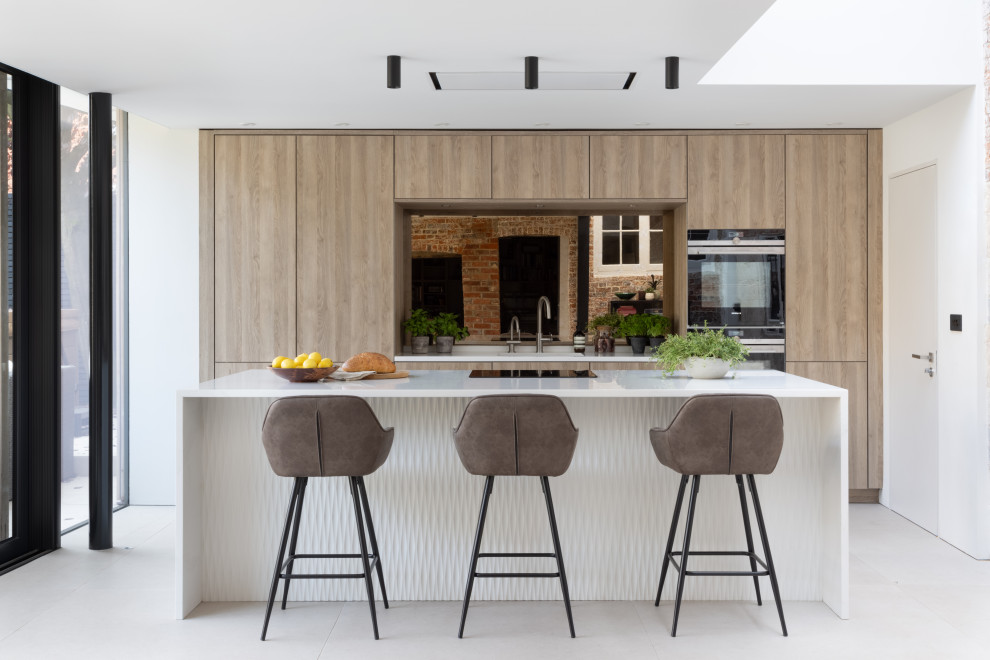 This is an example of a medium sized contemporary kitchen/diner in Hampshire with flat-panel cabinets, medium wood cabinets, composite countertops, mirror splashback, black appliances, an island and white worktops.