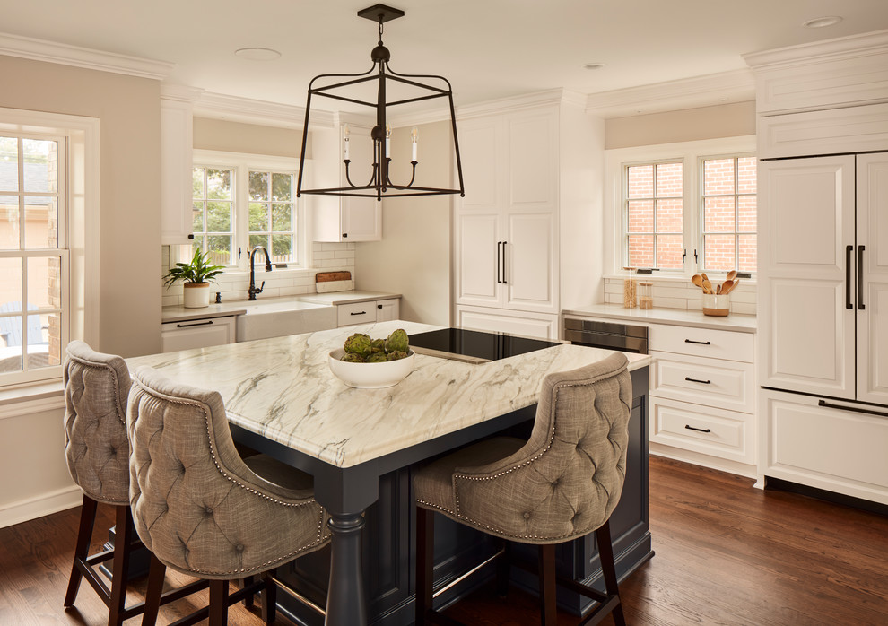 Traditional l-shaped kitchen in Milwaukee with a farmhouse sink, raised-panel cabinets, white cabinets, white splashback, subway tile splashback, panelled appliances, medium hardwood floors, with island, grey benchtop and marble benchtops.