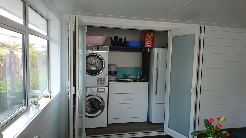 Photo of a modern single-wall laundry cupboard in Auckland with a drop-in sink, beaded inset cabinets, white cabinets, stainless steel benchtops, white walls, light hardwood floors, a stacked washer and dryer, brown floor and grey benchtop.