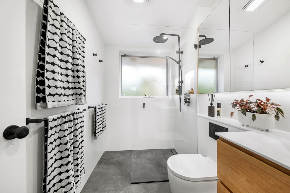 Свежая идея для дизайна: узкая и длинная главная ванная комната среднего размера в современном стиле с светлыми деревянными фасадами, открытым душем, унитазом-моноблоком, белой плиткой, керамической плиткой, белыми стенами, полом из керамической плитки, монолитной раковиной, столешницей из искусственного камня, серым полом, открытым душем, белой столешницей, тумбой под одну раковину, подвесной тумбой, сводчатым потолком и плоскими фасадами - отличное фото интерьера