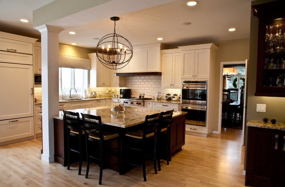 Example of a large transitional l-shaped light wood floor and brown floor open concept kitchen design with an undermount sink, shaker cabinets, white cabinets, granite countertops, white backsplash, subway tile backsplash, paneled appliances and an island