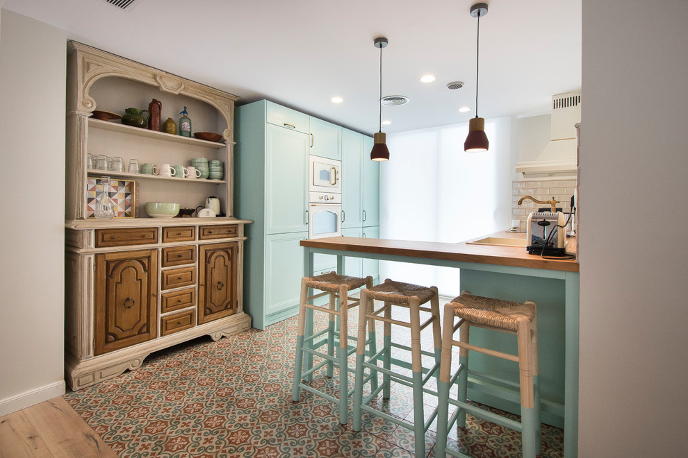 This is an example of a mid-sized mediterranean l-shaped kitchen in Other with turquoise cabinets, wood benchtops, ceramic floors, a peninsula, a drop-in sink, white splashback, multi-coloured floor and beige benchtop.