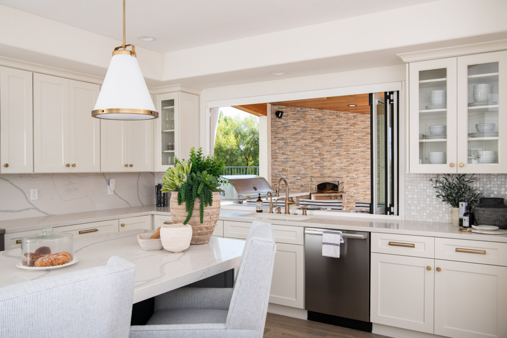 This is an example of a classic u-shaped open plan kitchen in Orange County with a submerged sink, shaker cabinets, white cabinets, engineered stone countertops, white splashback, engineered quartz splashback, stainless steel appliances, porcelain flooring, an island, brown floors and white worktops.