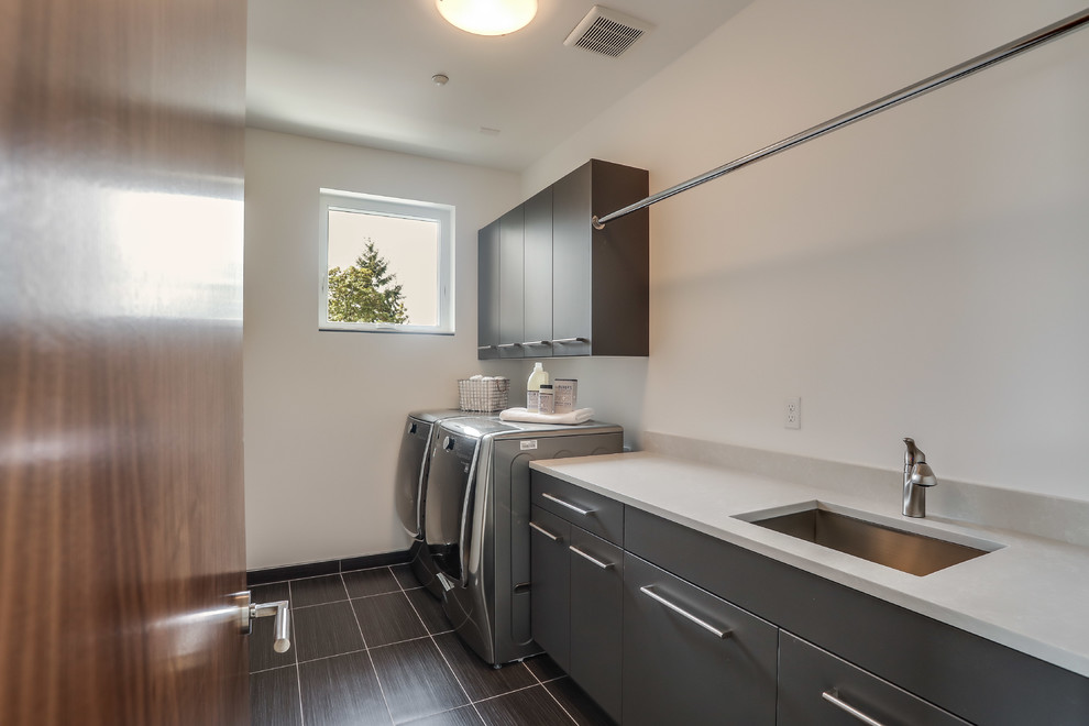 Design ideas for a mid-sized contemporary galley dedicated laundry room in Seattle with an undermount sink, flat-panel cabinets, grey cabinets, solid surface benchtops, white walls, ceramic floors, a side-by-side washer and dryer and grey floor.