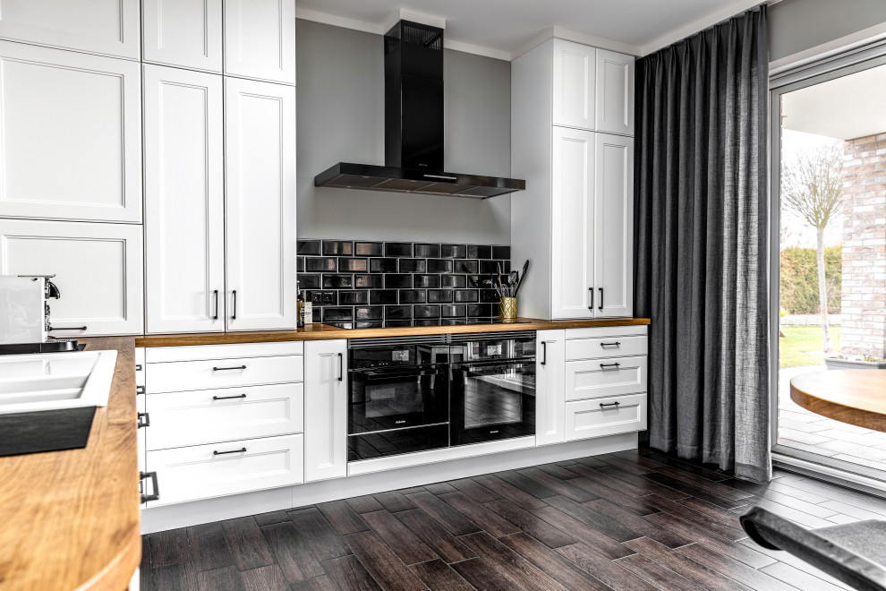 Mid-sized industrial l-shaped open plan kitchen in Berlin with a double-bowl sink, beaded inset cabinets, white cabinets, wood benchtops, black splashback, ceramic splashback, black appliances, porcelain floors, a peninsula, brown floor and brown benchtop.