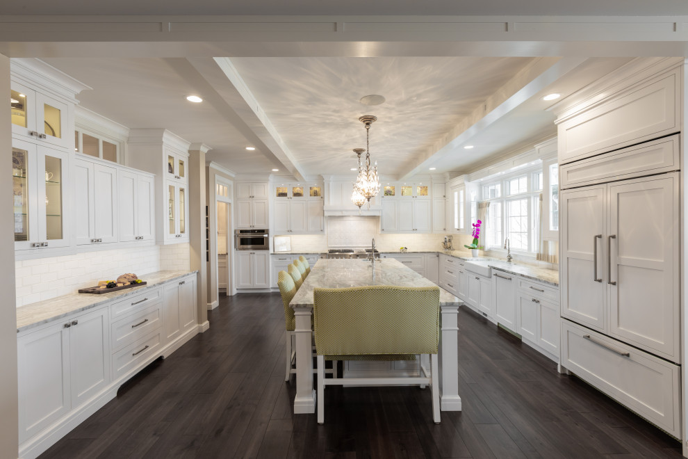 Example of a classic u-shaped dark wood floor and brown floor enclosed kitchen design in Other with a farmhouse sink, shaker cabinets, white cabinets, white backsplash, subway tile backsplash, paneled appliances and an island