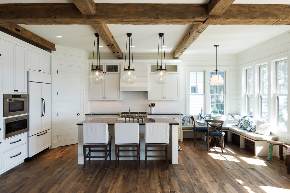 Large beach style l-shaped eat-in kitchen in Minneapolis with a farmhouse sink, shaker cabinets, white cabinets, granite benchtops, white splashback, subway tile splashback, panelled appliances, medium hardwood floors, with island and brown floor.
