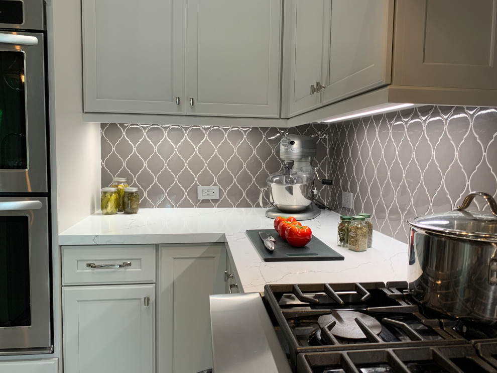 Example of a transitional l-shaped porcelain tile and multicolored floor kitchen design in Houston with a farmhouse sink, recessed-panel cabinets, white cabinets, quartz countertops, gray backsplash, ceramic backsplash, stainless steel appliances, an island and white countertops