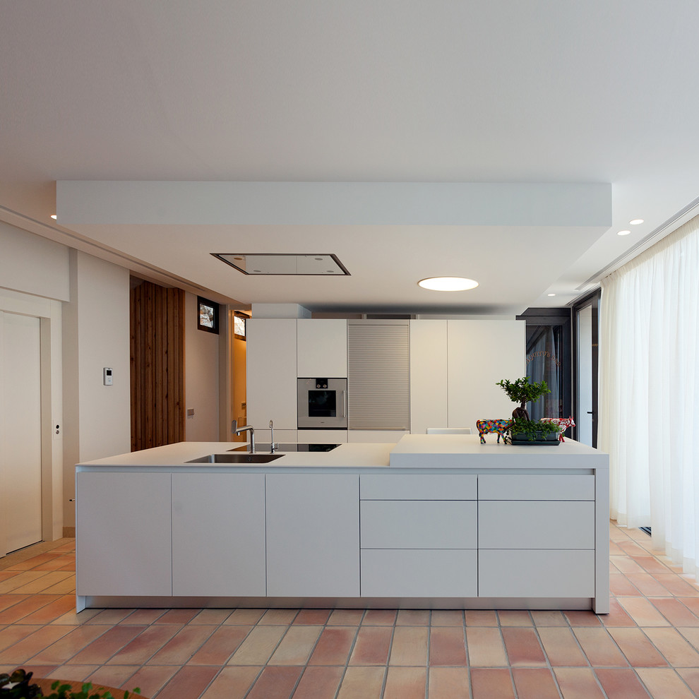 This is an example of a contemporary single-wall open plan kitchen in Valencia with flat-panel cabinets, white cabinets, with island, white benchtop, a drop-in sink, terra-cotta floors and red floor.