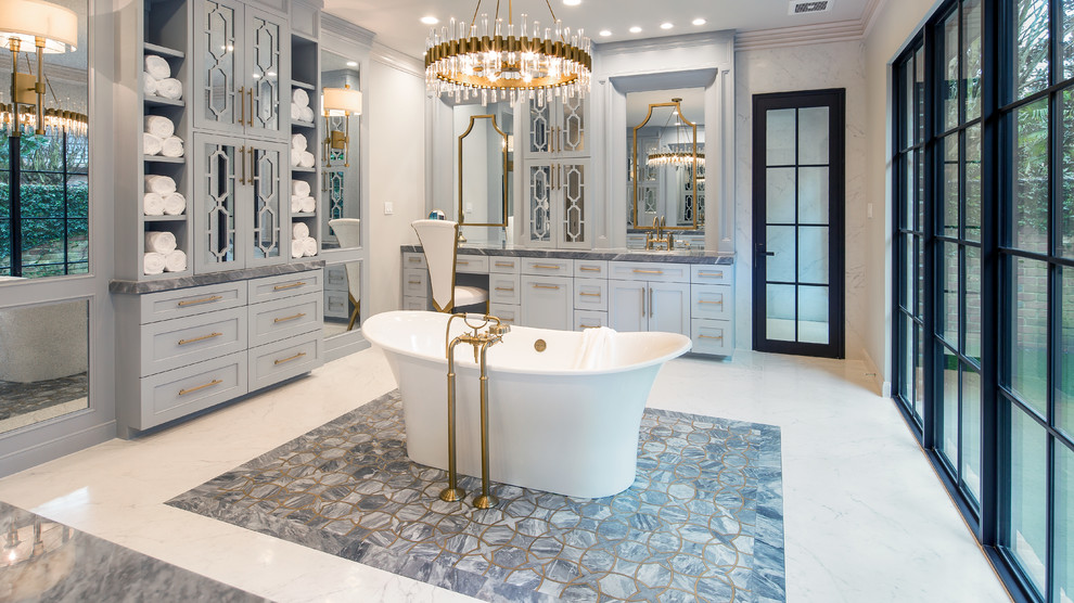 Photo of an expansive transitional master bathroom in Houston with furniture-like cabinets, grey cabinets, a freestanding tub, a curbless shower, a wall-mount toilet, white tile, porcelain tile, white walls, porcelain floors, a drop-in sink, marble benchtops, white floor and a hinged shower door.