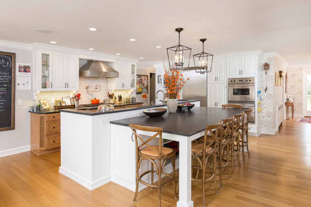 This is an example of a country l-shaped kitchen in Philadelphia with white cabinets, raised-panel cabinets, grey splashback, subway tile splashback, stainless steel appliances, medium hardwood floors, with island, brown floor and black benchtop.