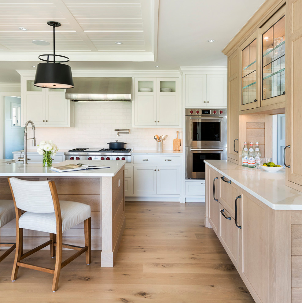 Photo of a beach style l-shaped kitchen in Providence with a farmhouse sink, white cabinets, white splashback, subway tile splashback, stainless steel appliances, light hardwood floors, with island, beige floor, white benchtop, timber and recessed.
