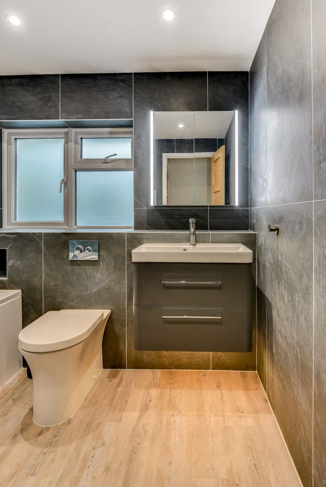 Exempel på ett mellanstort modernt badrum för barn, med släta luckor, grå skåp, ett badkar i en alkov, en dusch/badkar-kombination, en toalettstol med hel cisternkåpa, grå kakel, keramikplattor, grå väggar, ljust trägolv, ett nedsänkt handfat, brunt golv och dusch med gångjärnsdörr