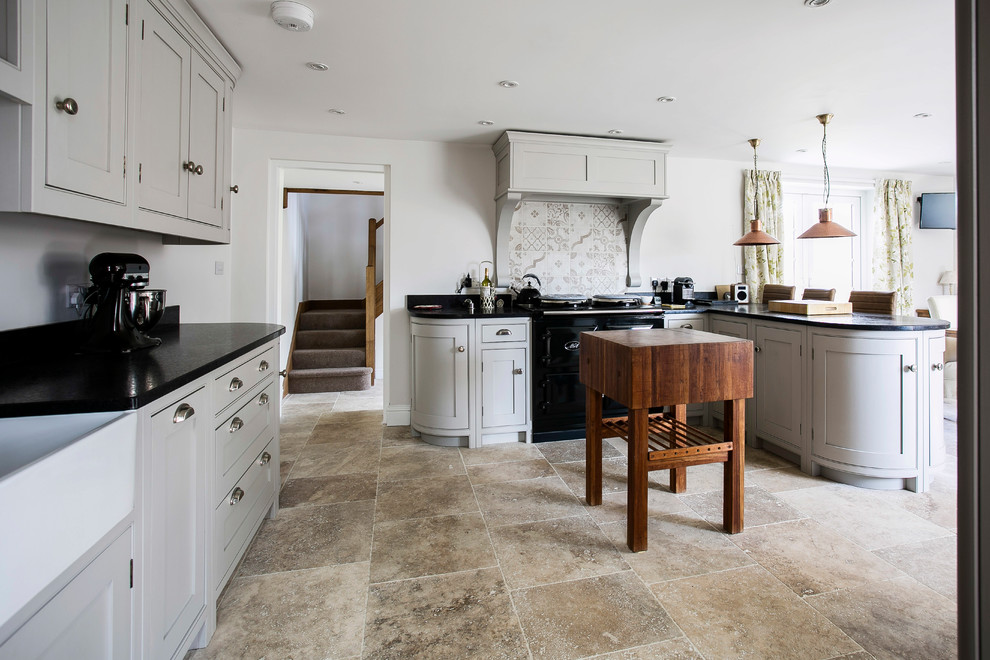 Photo of a mid-sized traditional u-shaped open plan kitchen in Kent with a farmhouse sink, shaker cabinets, grey cabinets, granite benchtops, brown splashback, ceramic splashback, black appliances, limestone floors, with island, beige floor and black benchtop.