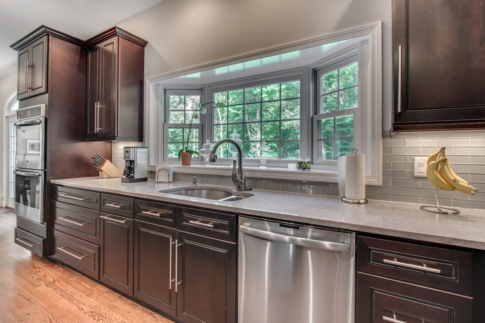 Eat-in kitchen - large transitional galley light wood floor and brown floor eat-in kitchen idea in Raleigh with an undermount sink, raised-panel cabinets, dark wood cabinets, quartz countertops, gray backsplash, glass tile backsplash, stainless steel appliances, an island and gray countertops