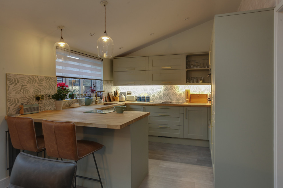 Photo of a medium sized contemporary u-shaped kitchen/diner in Essex with an integrated sink, shaker cabinets, green cabinets, wood worktops, porcelain splashback, black appliances, laminate floors and a breakfast bar.