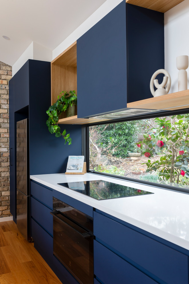 This is an example of a medium sized contemporary galley open plan kitchen in Canberra - Queanbeyan with a submerged sink, all styles of cabinet, blue cabinets, engineered stone countertops, window splashback, black appliances, medium hardwood flooring, an island and white worktops.