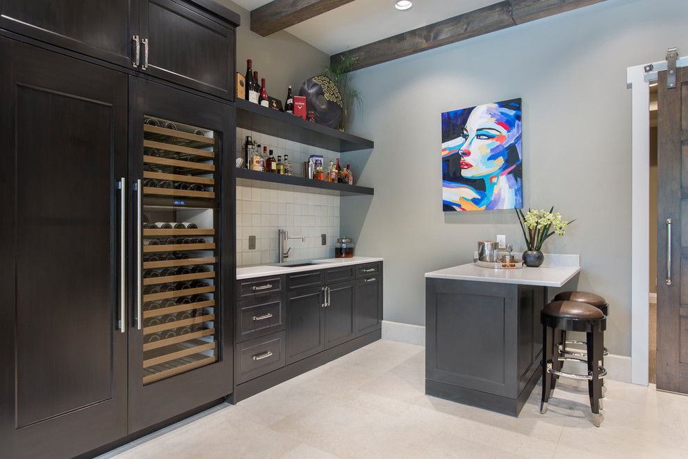 Photo of a transitional seated home bar in Dallas with recessed-panel cabinets, grey cabinets, quartz benchtops, beige splashback, ceramic splashback, limestone floors, beige floor and white benchtop.