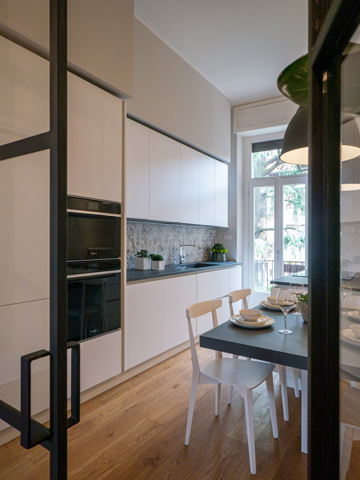 Photo of a large contemporary galley separate kitchen in Milan with an integrated sink, flat-panel cabinets, white cabinets, solid surface benchtops, multi-coloured splashback, porcelain splashback, black appliances, light hardwood floors, with island, grey benchtop and recessed.