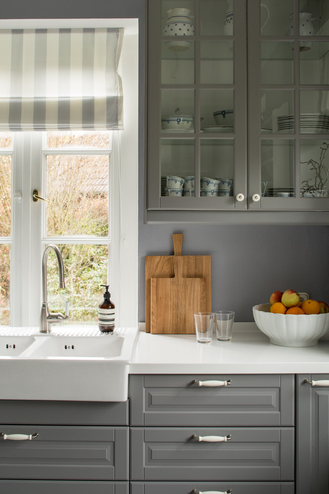 Country kitchen in Hamburg with a farmhouse sink, raised-panel cabinets, grey cabinets, grey splashback and white benchtop.