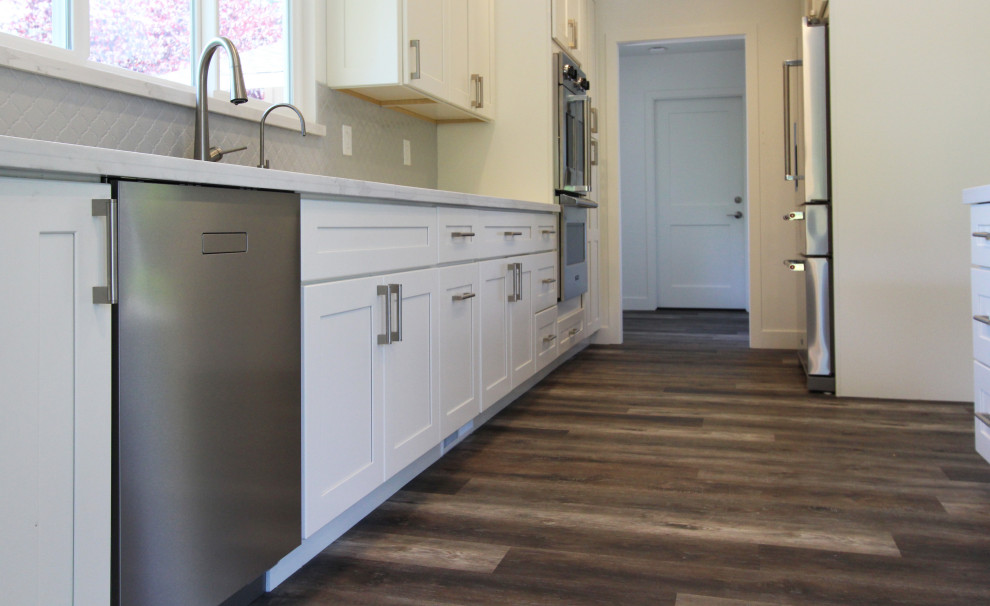Large transitional single-wall vinyl floor and brown floor open concept kitchen photo with an undermount sink, shaker cabinets, white cabinets, quartz countertops, gray backsplash, ceramic backsplash, stainless steel appliances, an island and white countertops