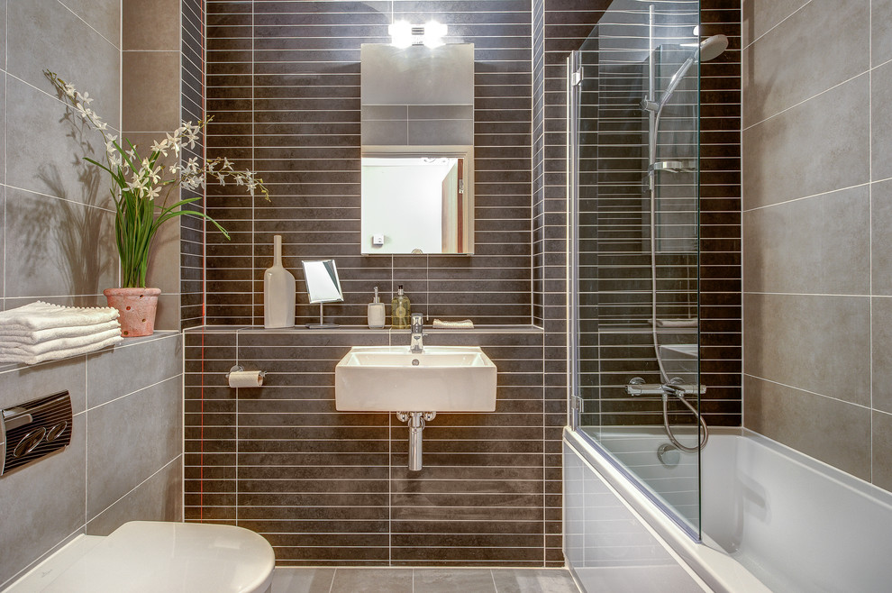 Small contemporary 3/4 bathroom in Devon with a wall-mount sink, tile benchtops, an alcove tub, a shower/bathtub combo, a one-piece toilet, brown tile, ceramic tile, beige walls and ceramic floors.