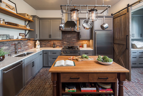Rustic-industrial kitchen with gray shaker kitchen cabinet fronts, brick backsplash and floor, butcher-block island, hanging pot-rack lights, stainless refrigerator, and a sliding barn door.