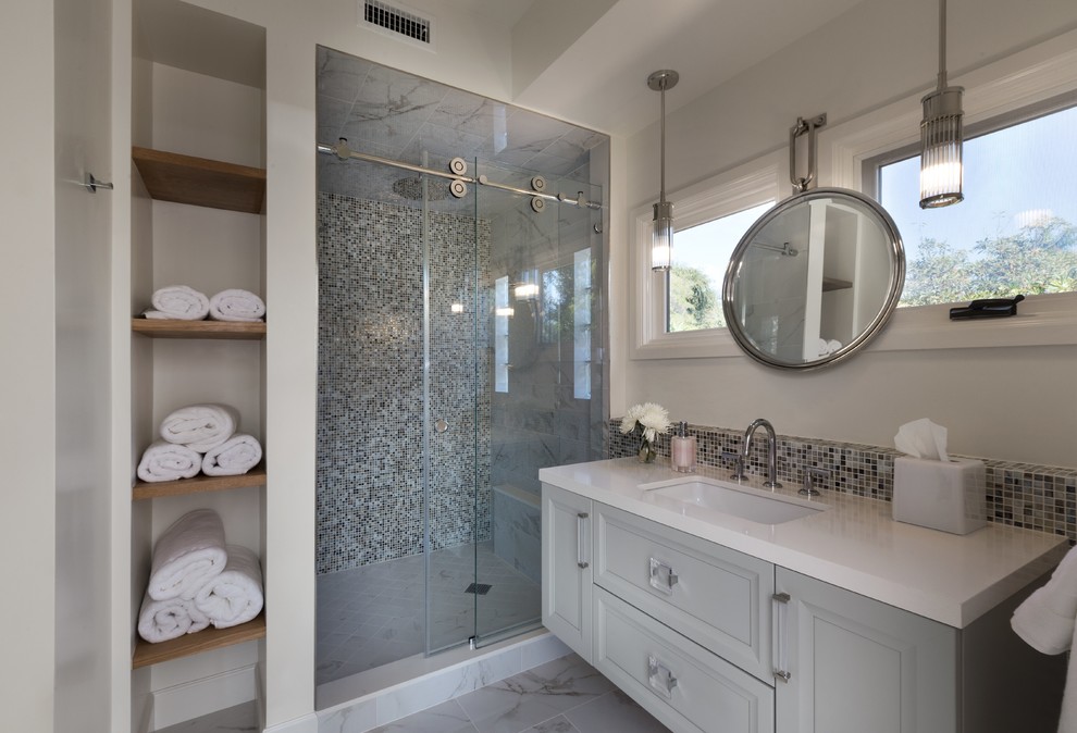 Design ideas for a mid-sized transitional 3/4 bathroom in Los Angeles with grey cabinets, gray tile, mosaic tile, white walls, an undermount sink, white floor, a sliding shower screen, white benchtops, raised-panel cabinets, an alcove shower, marble floors and engineered quartz benchtops.
