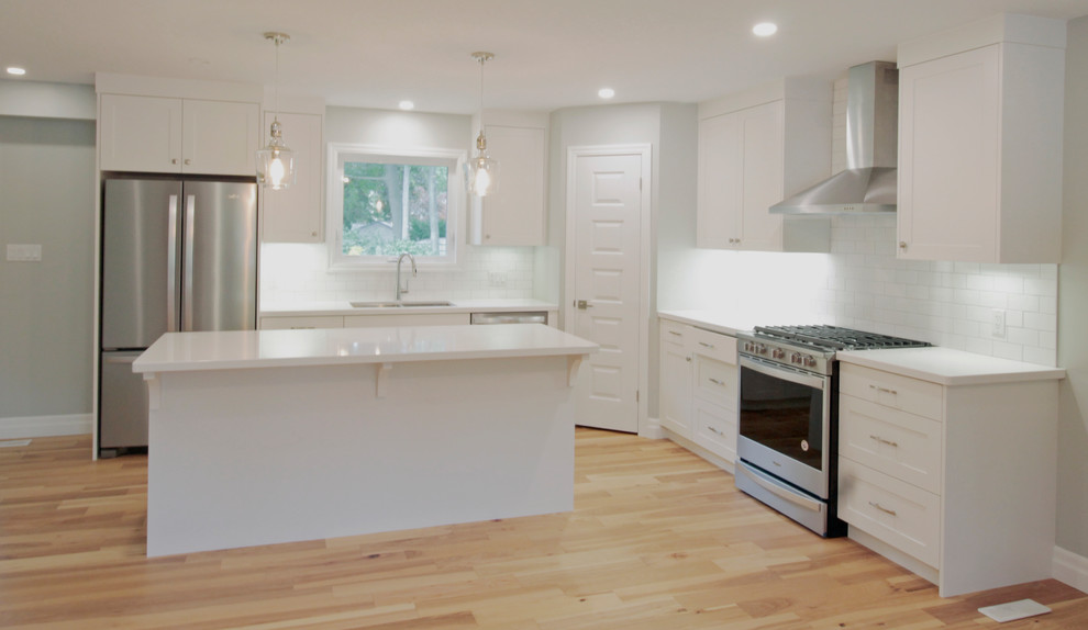 Example of a mid-sized transitional l-shaped light wood floor eat-in kitchen design with a double-bowl sink, shaker cabinets, white cabinets, solid surface countertops, white backsplash, ceramic backsplash, stainless steel appliances, an island and white countertops